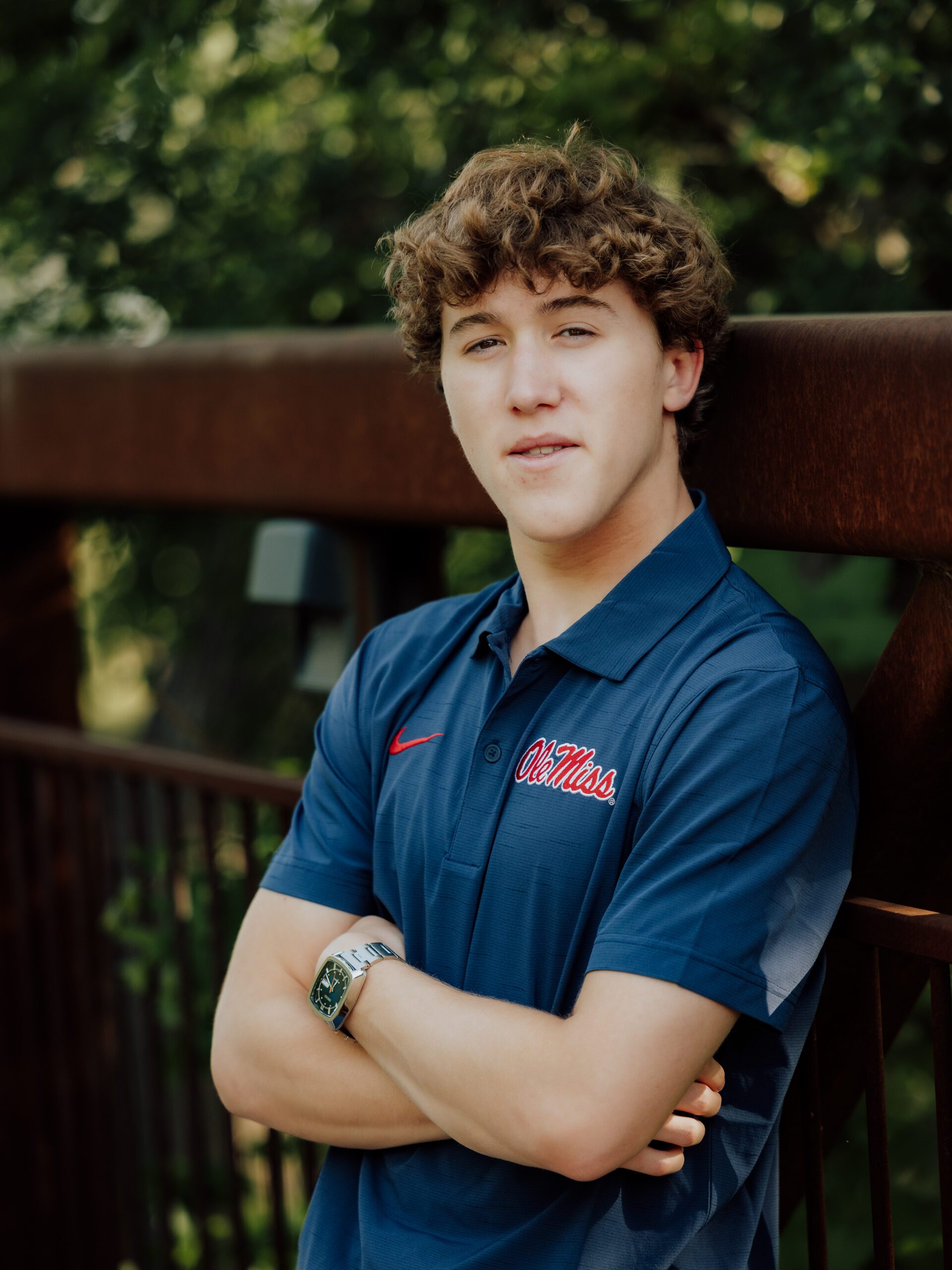 High school senior boy in Ole Miss polo with arms crossed, leaning against a wooden railing with lush green bokeh background — classic senior portrait by William Bichara Photography, Dallas TX