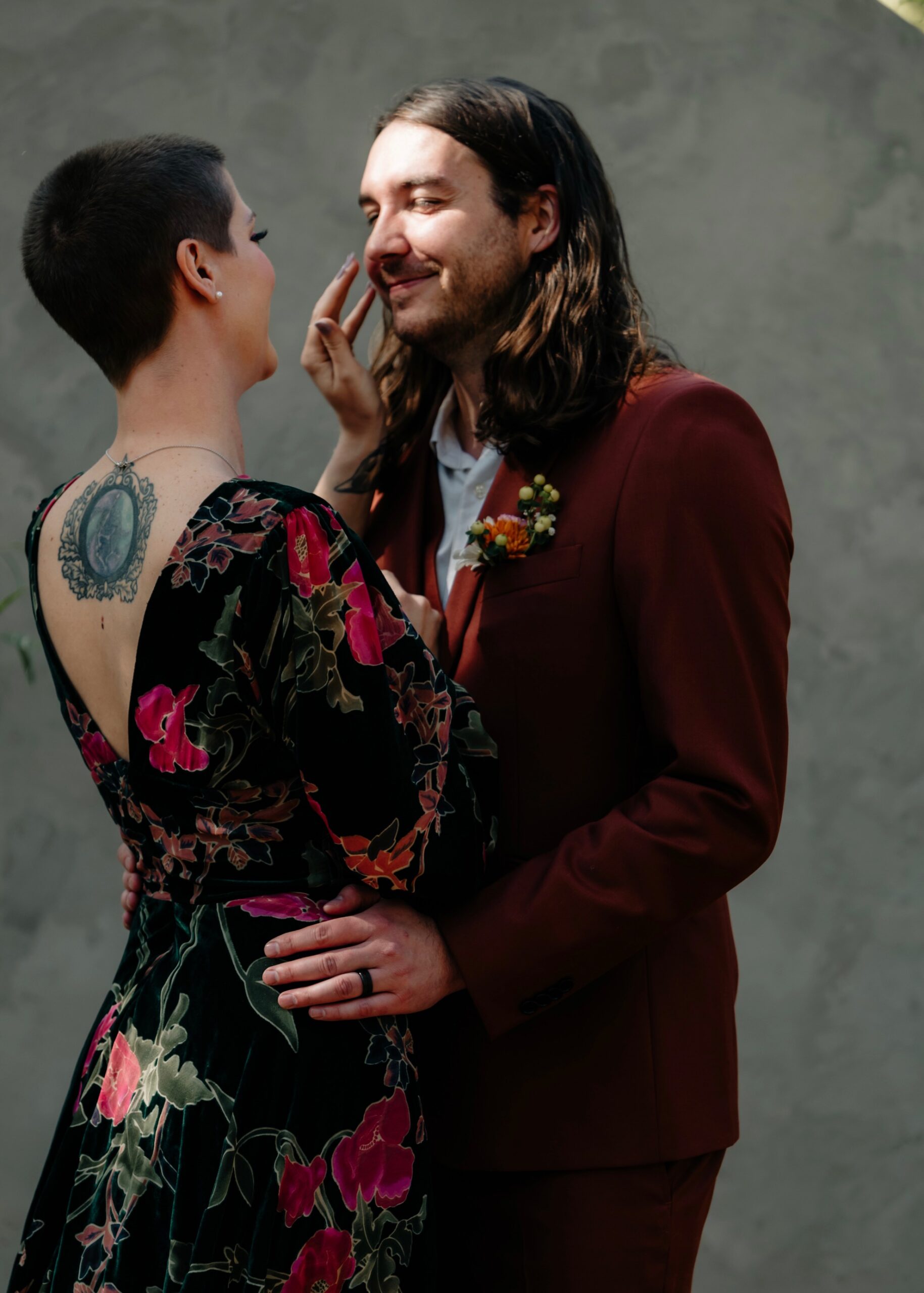 Bride in a floral gown playfully touches her partner's face, both smiling, in front of a stone archway at an outdoor ceremony