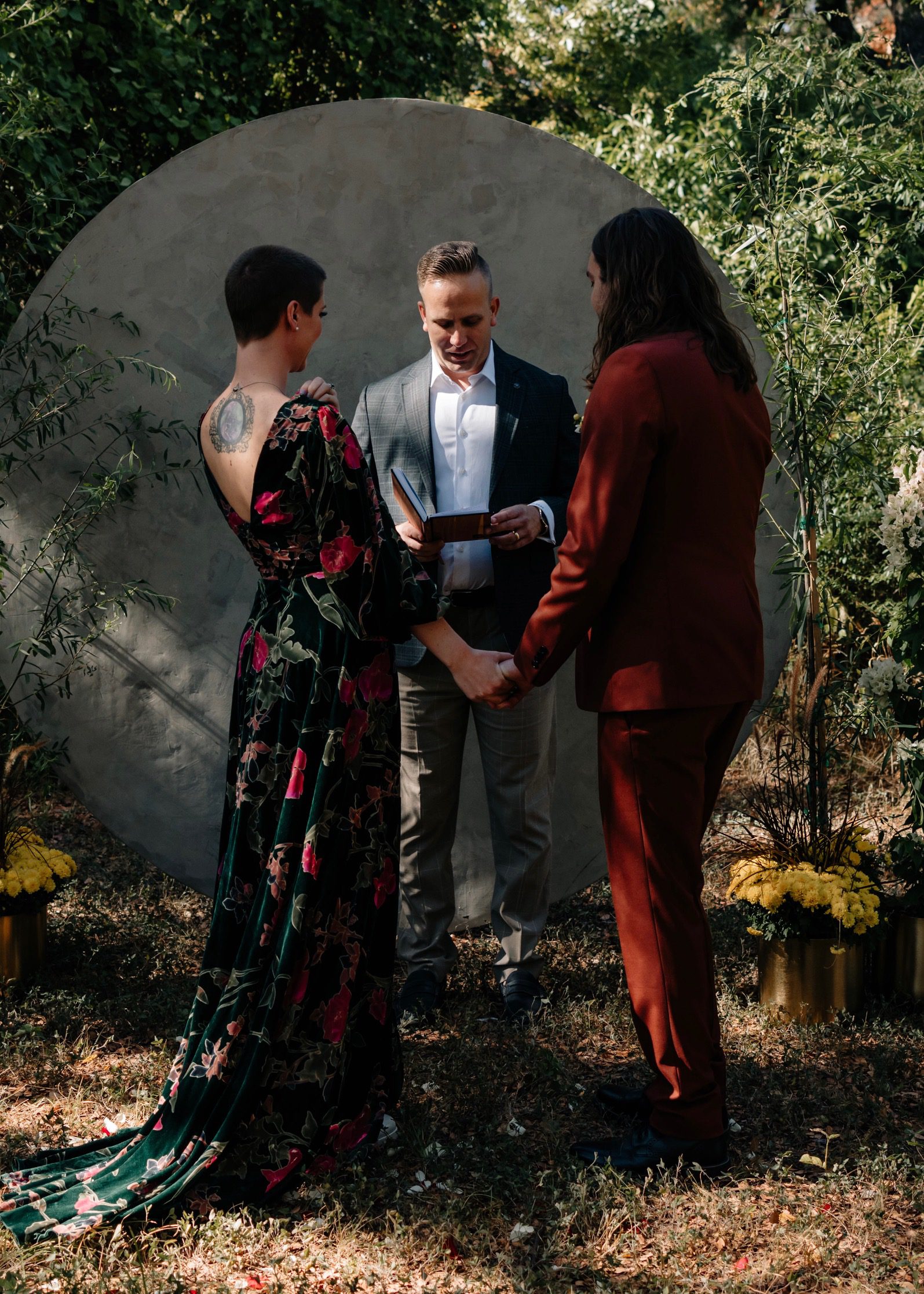 Officiant joins hands of bride and groom during an outdoor elopement ceremony beneath a stone arch surrounded by greenery