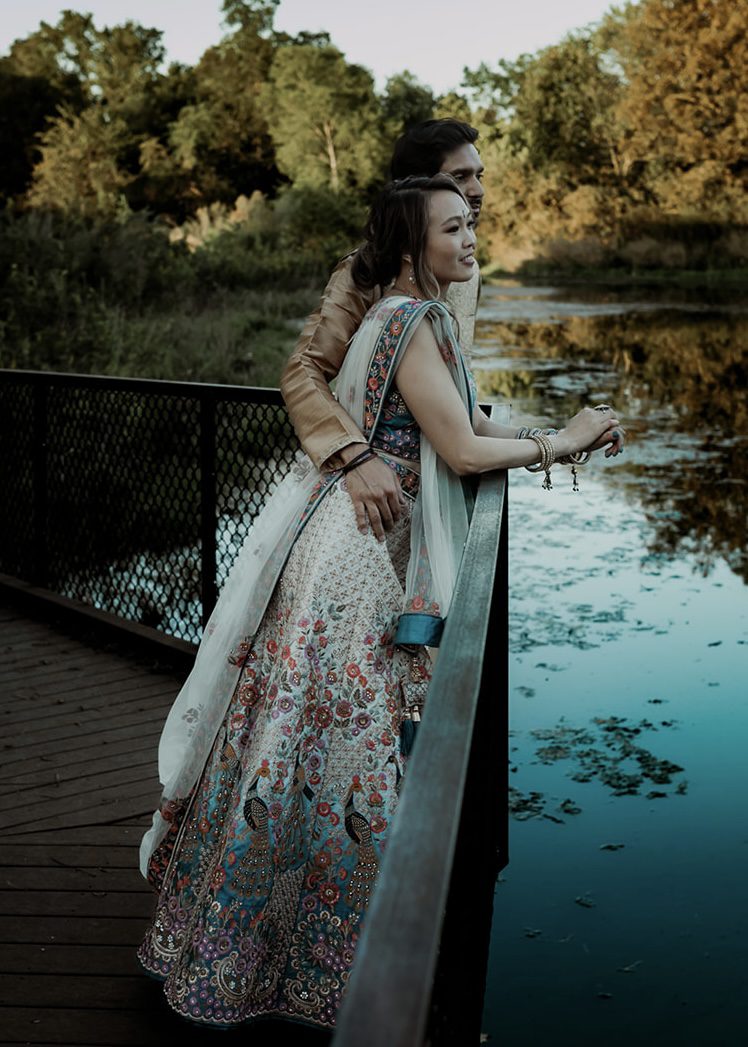 Bride in an embellished teal and ivory lehenga leaning against a railing overlooking a river, groom standing close behind her