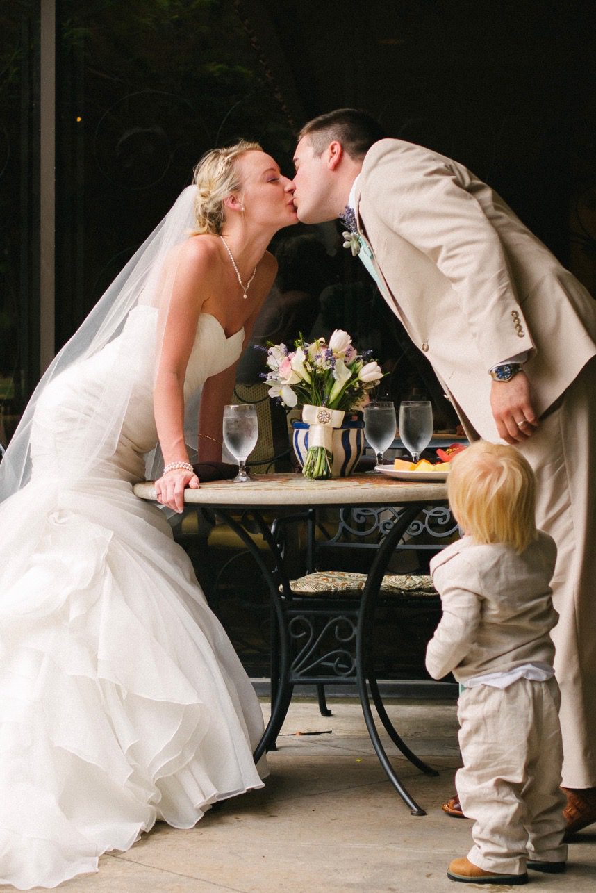 Bride and groom kiss over a reception table while a toddler in a linen suit stands nearby