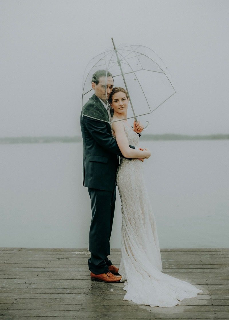 Bride and groom embrace under a clear umbrella on a foggy dock overlooking a lake, bride looking back over her shoulder